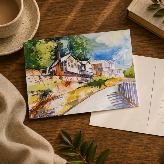 A watercolor postcard featuring a scenic house illustration on a wooden table, placed next to an empty white postcard envelope. The scene is styled with a cup of coffee, a book, and green leaves.