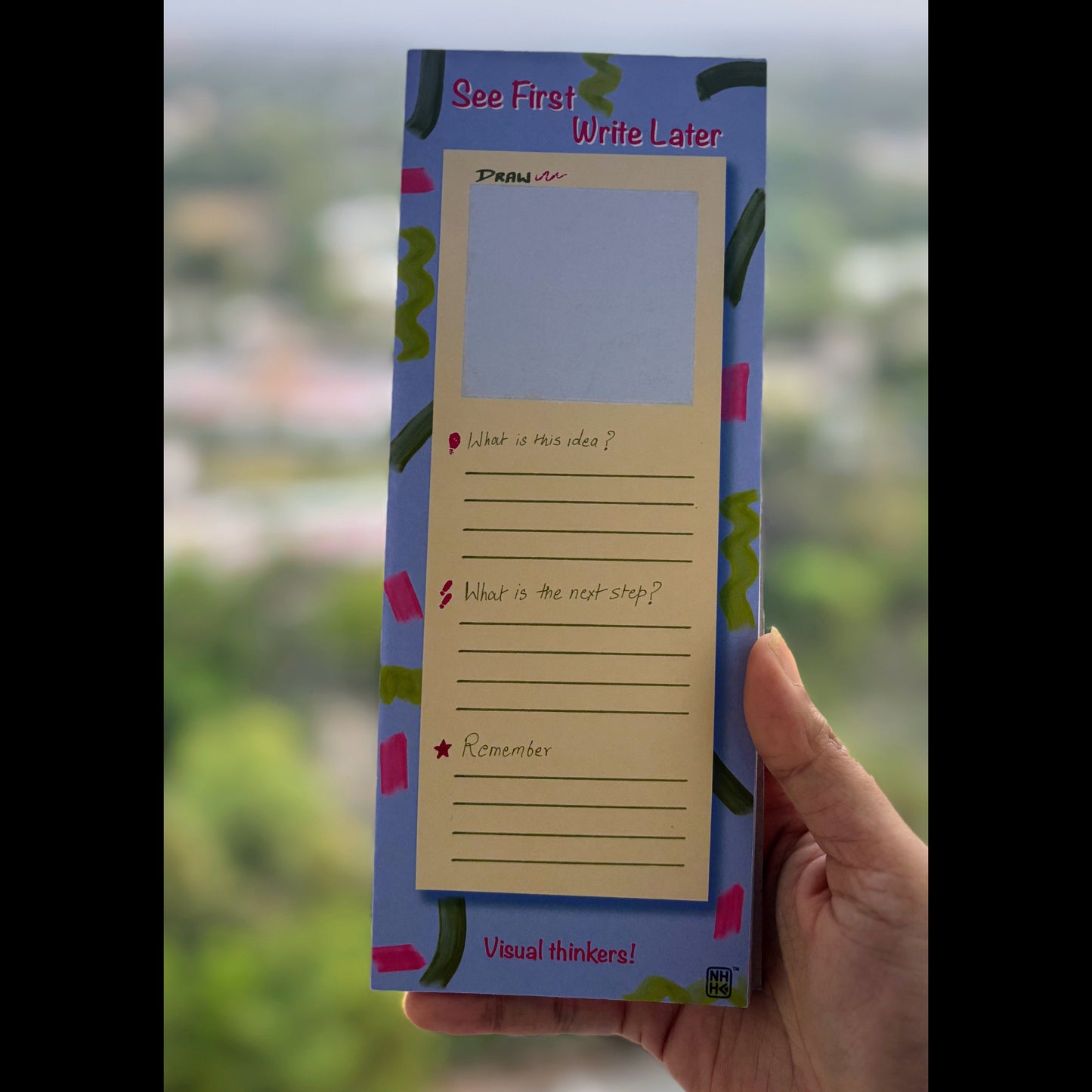 A hand holding a blue and pink 'See First Write Later' sticky note pad with a blank writing area and lined prompts, set against a blurred outdoor background.