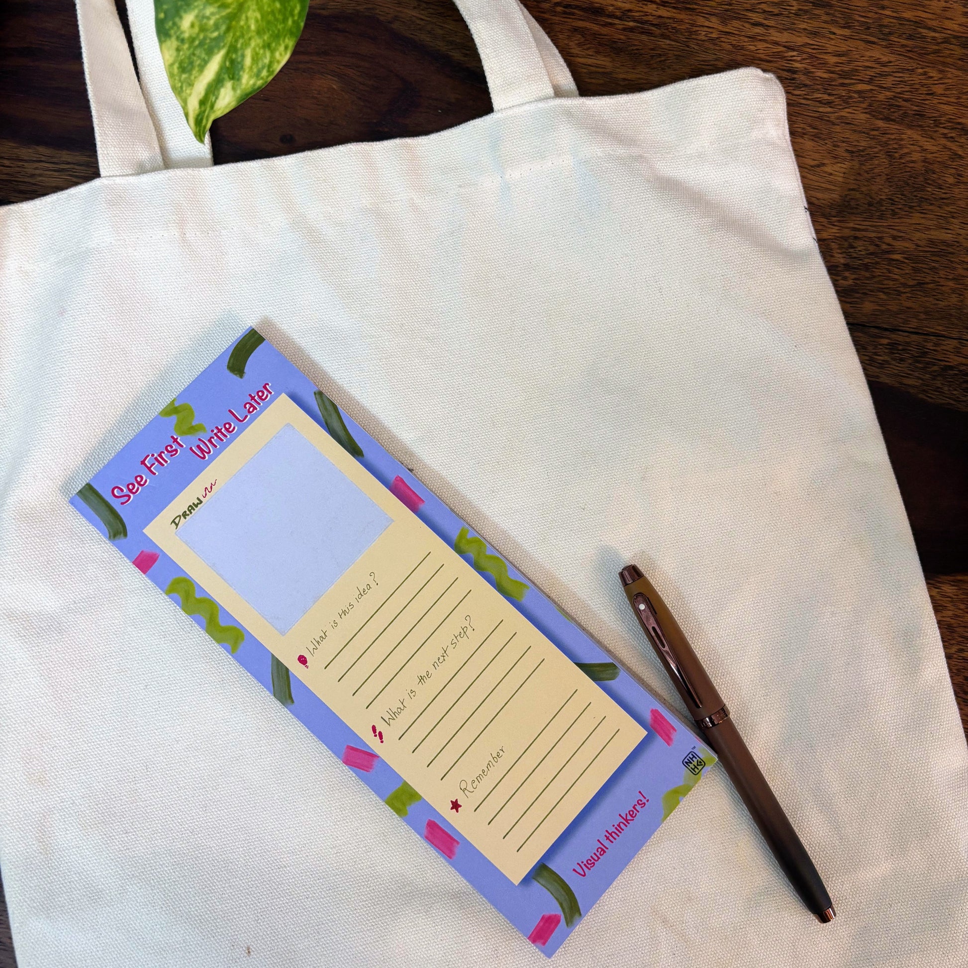 A white canvas tote bag laid flat on a dark wooden table, styled with a 'Sun Free Week Later' notepad and a brown pen.