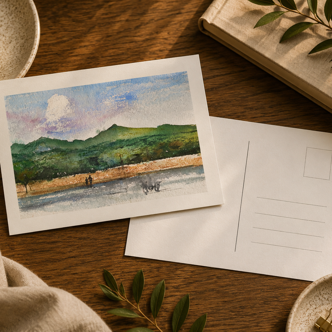 A watercolor postcard featuring a scenic landscape with green hills, a blue sky, and a river, displayed next to its blank white back on a wooden table with lifestyle props like a book, cup of coffee, and linen.
